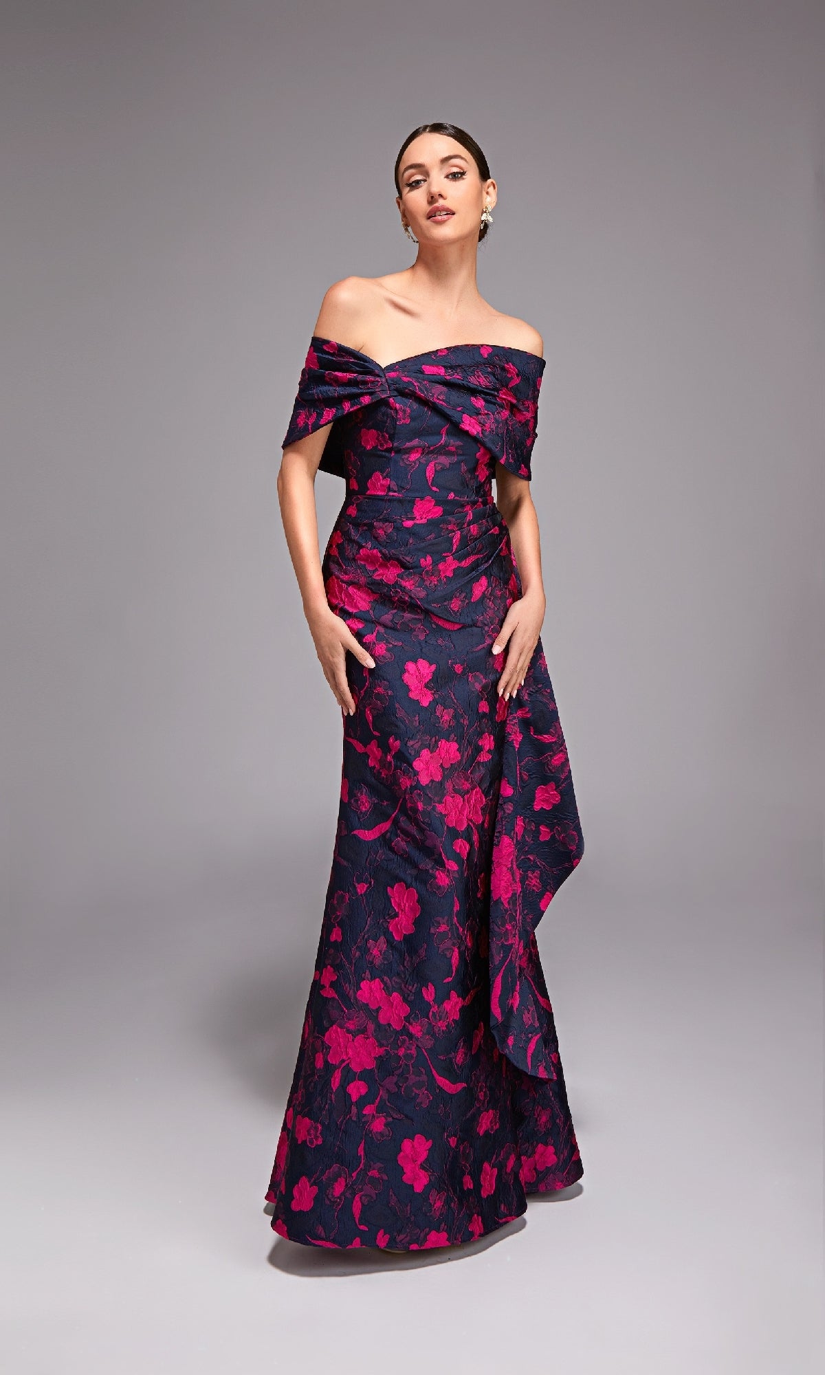 Woman in the Alyce 27832 long formal dress, a floral jacquard gown with bright pink blooms on a dark base, off-the-shoulder and floor-length, standing against a plain gray background. Ideal for mother-of-the-bride.
